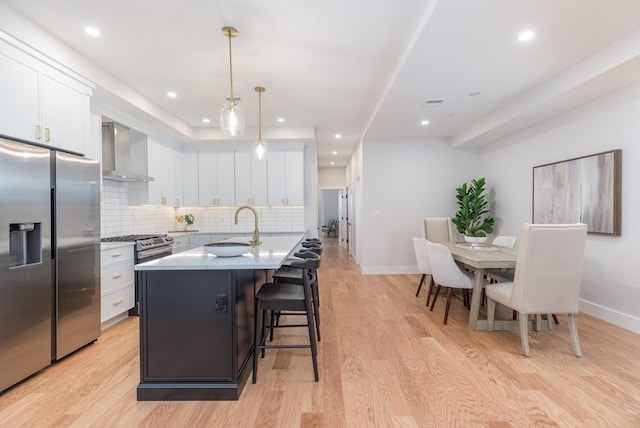 kitchen featuring wall chimney exhaust hood, hanging light fixtures, stainless steel appliances, a kitchen island with sink, and white cabinets