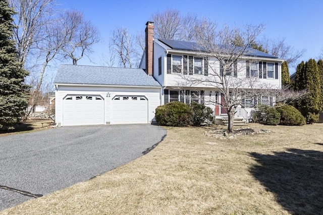 colonial-style house with a front lawn, roof mounted solar panels, a chimney, a garage, and driveway