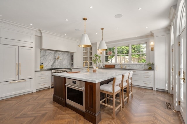 kitchen featuring white cabinets, light stone countertops, an island with sink, stainless steel oven, and pendant lighting