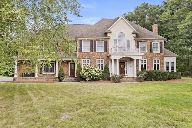 colonial-style house featuring a balcony, brick siding, roof with shingles, a front lawn, and a chimney