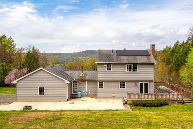 back of house featuring a deck, a patio area, roof mounted solar panels, a chimney, and a lawn