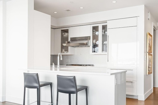 kitchen featuring visible vents, a breakfast bar, glass insert cabinets, wall chimney range hood, and tasteful backsplash