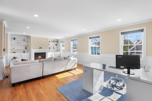 living area featuring a warm lit fireplace, light wood finished floors, wainscoting, and built in shelves