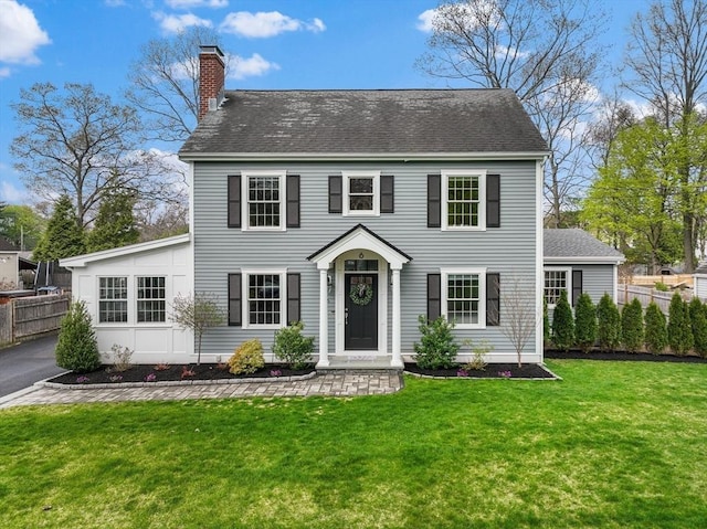 colonial home with roof with shingles, fence, a front yard, and a chimney
