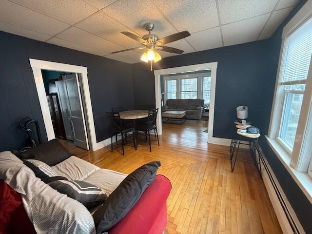 living room with ceiling fan, a paneled ceiling, wood-type flooring, and a baseboard heating unit