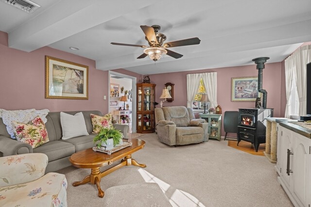 carpeted living room featuring a wood stove, ceiling fan, and beam ceiling