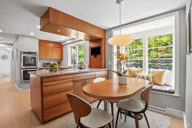 kitchen with double oven, brown cabinets, light wood-style flooring, light stone countertops, and pendant lighting
