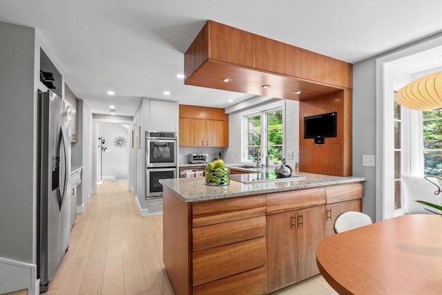 kitchen with appliances with stainless steel finishes, a peninsula, light stone counters, a sink, and light wood-style flooring