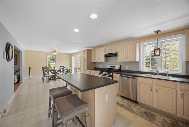 kitchen with light brown cabinetry, dark countertops, under cabinet range hood, a sink, and stainless steel appliances