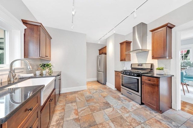 kitchen with appliances with stainless steel finishes, wall chimney exhaust hood, track lighting, dark stone counters, and baseboards