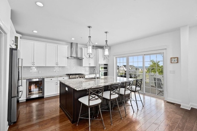 kitchen with sink, wall chimney range hood, pendant lighting, a center island with sink, and wine cooler