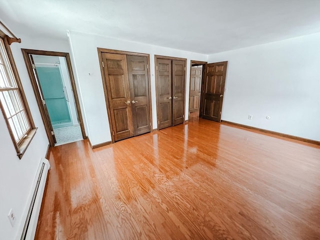 unfurnished bedroom featuring a baseboard heating unit, baseboards, two closets, and light wood-style floors