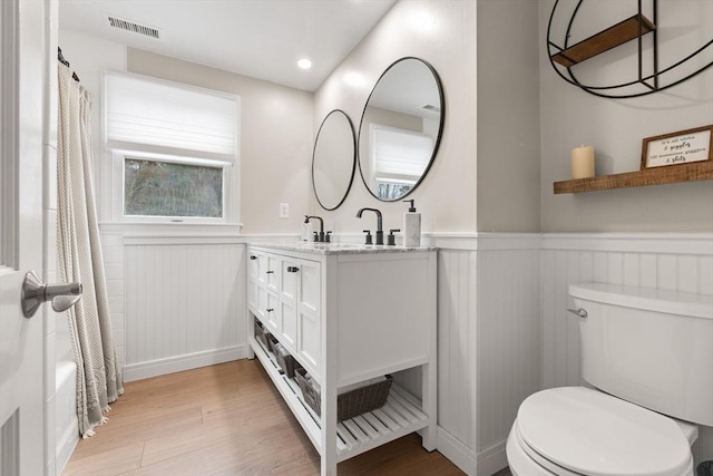full bath featuring double vanity, visible vents, wainscoting, wood finished floors, and toilet