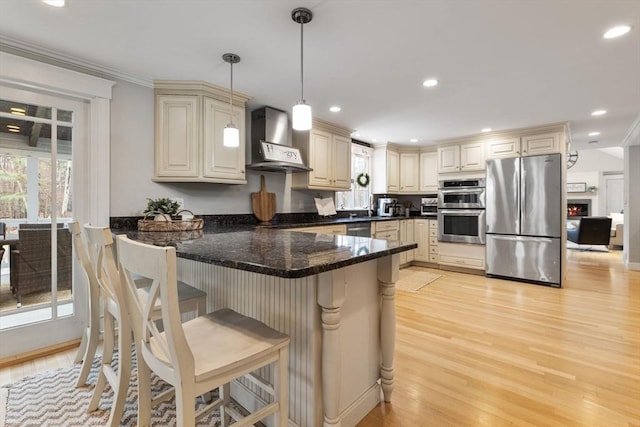 kitchen with a peninsula, light wood-style floors, wall chimney exhaust hood, cream cabinets, and appliances with stainless steel finishes