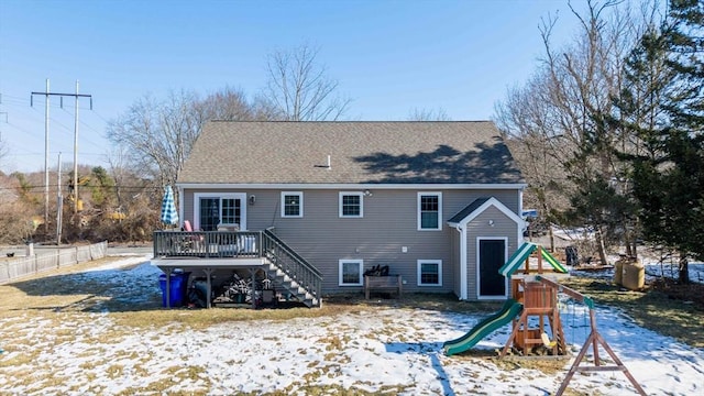 snow covered back of property featuring fence, a deck, a playground, and roof with shingles