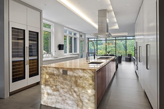 kitchen featuring light stone countertops, white cabinets, island exhaust hood, a kitchen island with sink, and sink