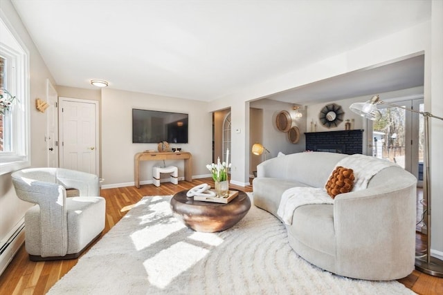 living room with a brick fireplace, a baseboard heating unit, baseboards, and wood finished floors