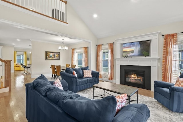 living room featuring a notable chandelier, crown molding, light wood-style flooring, a glass covered fireplace, and high vaulted ceiling