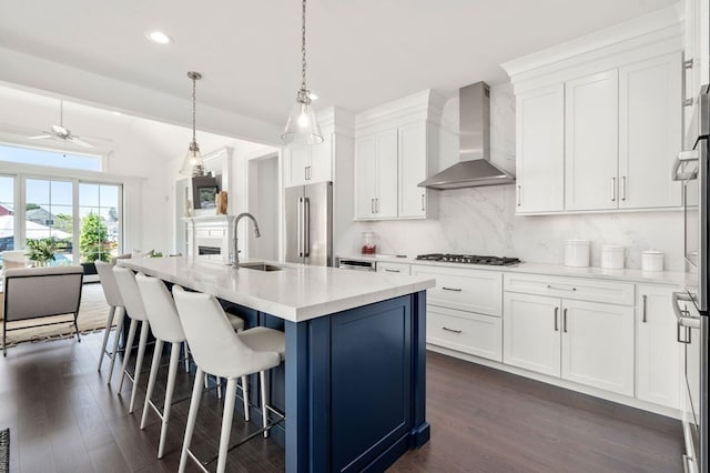 kitchen with sink, hanging light fixtures, wall chimney range hood, an island with sink, and appliances with stainless steel finishes