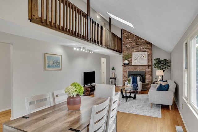 living room featuring light wood-style flooring, a brick fireplace, a skylight, high vaulted ceiling, and baseboards