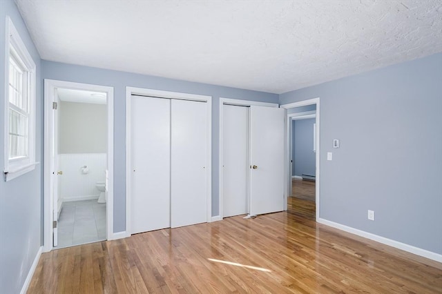 unfurnished bedroom featuring ensuite bath, a textured ceiling, light hardwood / wood-style floors, baseboard heating, and two closets