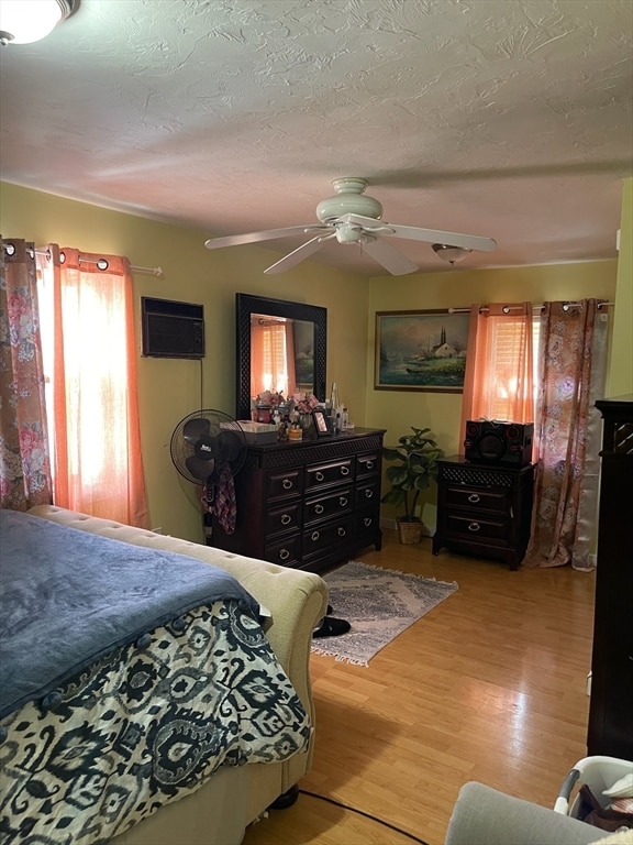 bedroom with a wall mounted air conditioner, a textured ceiling, light wood-type flooring, and ceiling fan