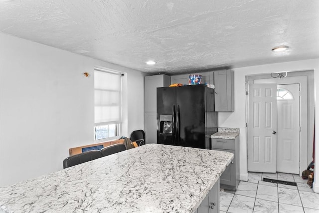 kitchen with light stone counters, marble finish floor, gray cabinets, black refrigerator with ice dispenser, and a textured ceiling