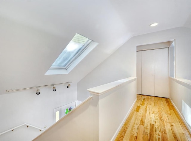 corridor featuring rail lighting, lofted ceiling with skylight, an upstairs landing, light wood-type flooring, and plenty of natural light