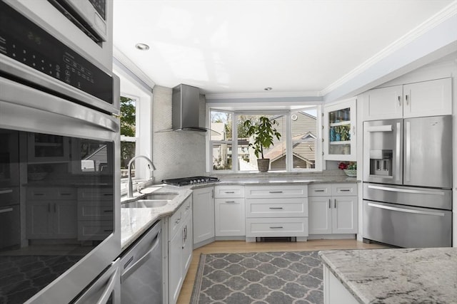 kitchen featuring wall chimney range hood, appliances with stainless steel finishes, a sink, light stone counters, and ornamental molding