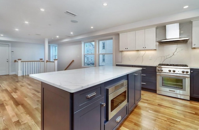 kitchen with wall chimney range hood, light wood-type flooring, appliances with stainless steel finishes, a kitchen island, and white cabinetry