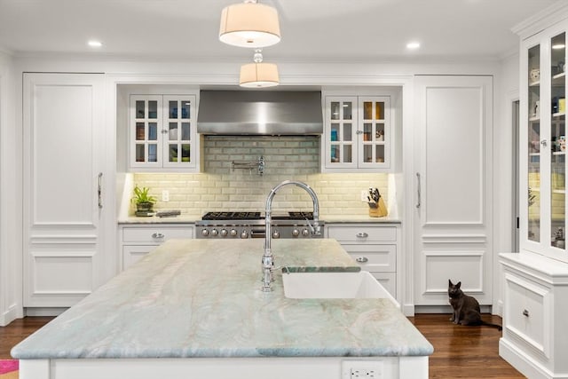 kitchen with wall chimney exhaust hood, a center island with sink, dark wood-type flooring, tasteful backsplash, and ornamental molding