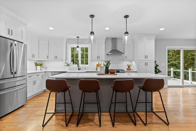 kitchen with wall chimney range hood, decorative light fixtures, white cabinets, a center island, and stainless steel refrigerator