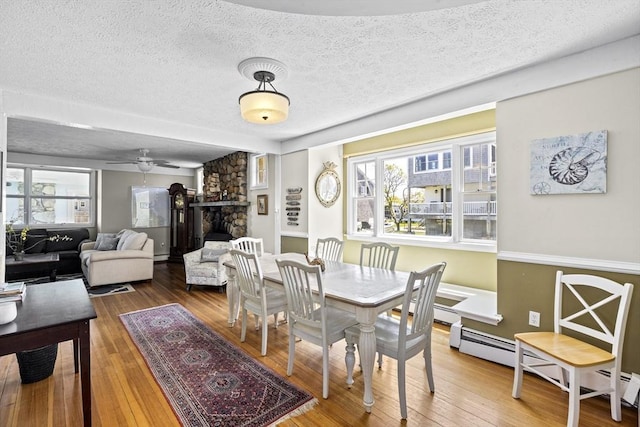 dining space with a baseboard heating unit, ceiling fan, a fireplace, wood-type flooring, and a textured ceiling