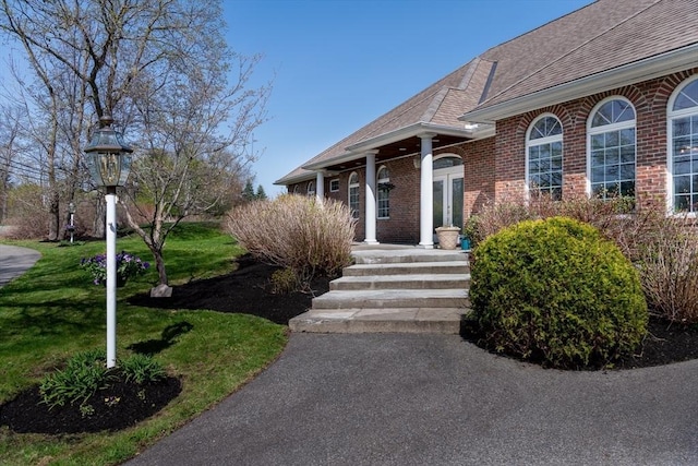 entrance to property featuring roof with shingles, a yard, and brick siding