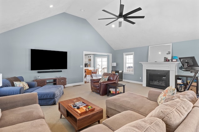 living area featuring baseboards, a ceiling fan, a glass covered fireplace, light colored carpet, and high vaulted ceiling