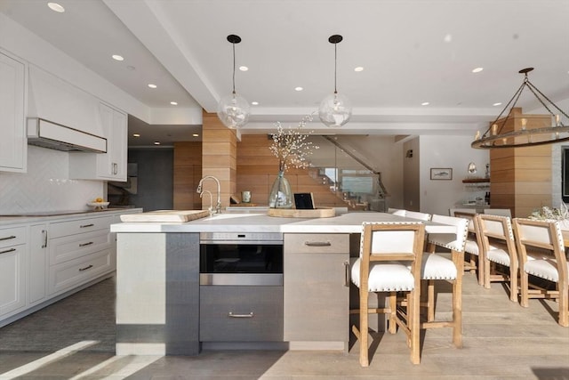 kitchen with stainless steel oven, white cabinets, a center island with sink, hanging light fixtures, and decorative backsplash