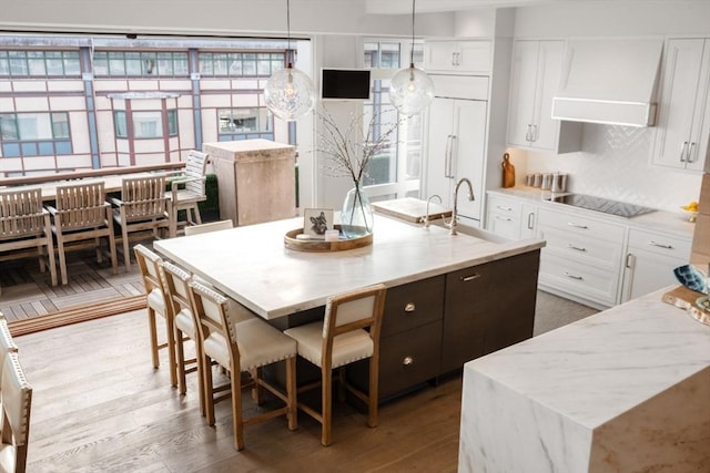 kitchen with black electric stovetop, custom exhaust hood, a kitchen island with sink, white cabinetry, and hanging light fixtures