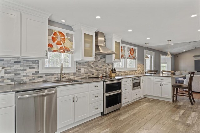 kitchen featuring a peninsula, a sink, stainless steel appliances, white cabinetry, and wall chimney exhaust hood