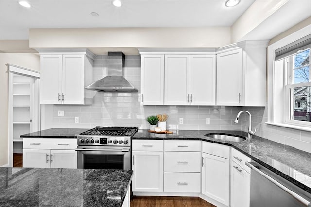 kitchen featuring sink, stainless steel appliances, wall chimney range hood, dark stone counters, and white cabinets