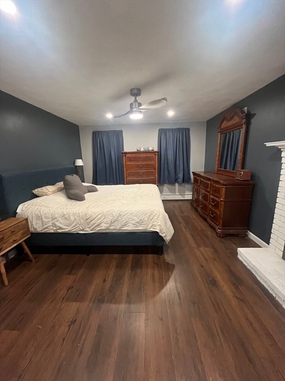 bedroom featuring dark wood-type flooring, baseboard heating, ceiling fan, and a brick fireplace