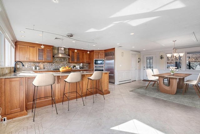 kitchen with sink, hanging light fixtures, a kitchen breakfast bar, built in appliances, and wall chimney exhaust hood