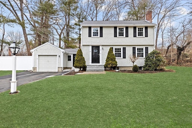 colonial-style house featuring a front yard, a chimney, an attached garage, fence, and aphalt driveway