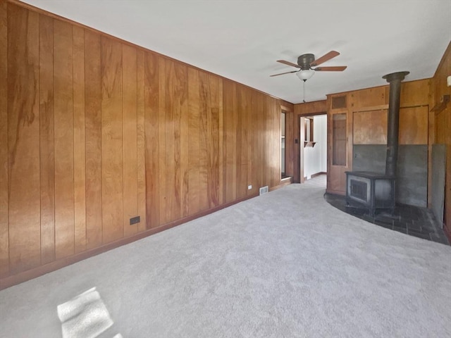 unfurnished living room with visible vents, a wood stove, ceiling fan, wooden walls, and carpet