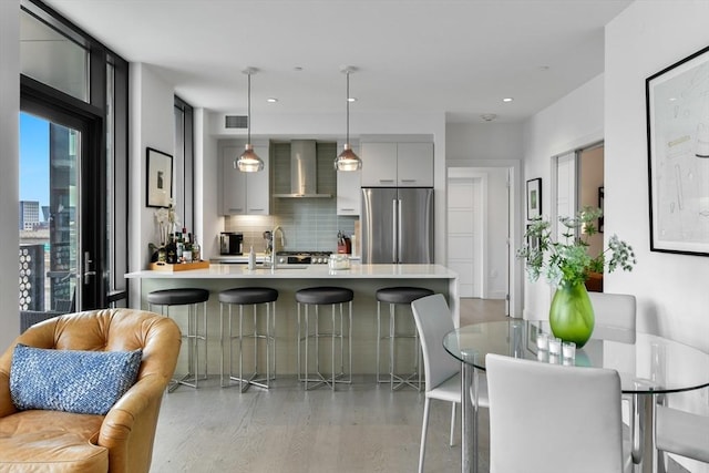 kitchen with freestanding refrigerator, wall chimney range hood, light countertops, a kitchen breakfast bar, and modern cabinets