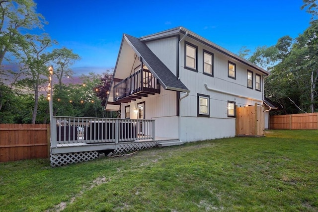 back of house at dusk with fence, a wooden deck, a lawn, a balcony, and roof with shingles