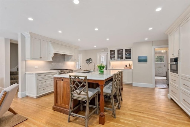 kitchen with a kitchen island with sink, custom exhaust hood, light countertops, stainless steel oven, and a sink