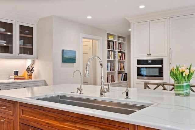 kitchen with light stone counters, decorative backsplash, wall oven, glass insert cabinets, and a sink