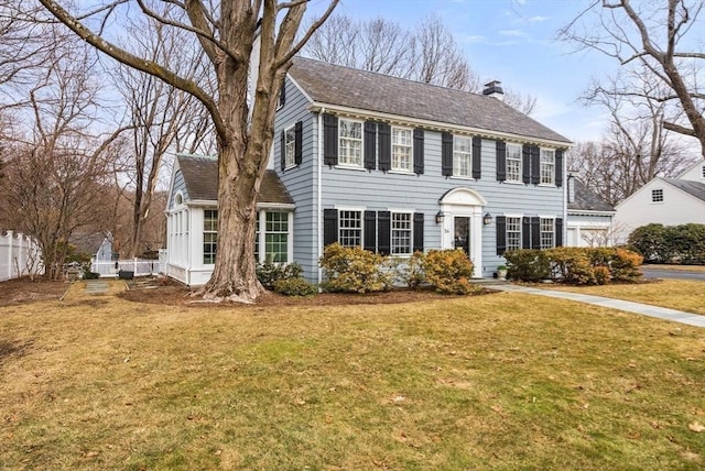 colonial-style house with a shingled roof, a chimney, fence, and a front yard