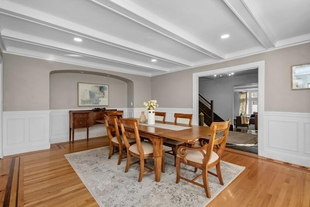 dining space with light wood-type flooring, arched walkways, beam ceiling, and wainscoting