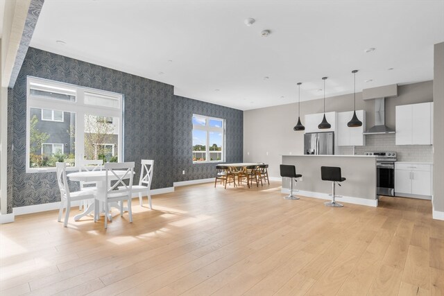 kitchen with wall chimney range hood, hanging light fixtures, appliances with stainless steel finishes, white cabinetry, and light wood-type flooring